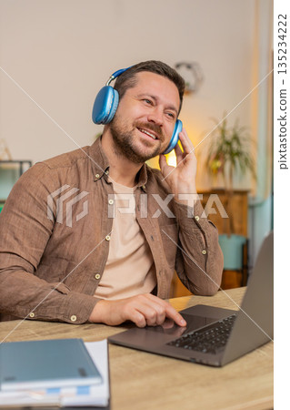 Happy Caucasian man using laptop while listening to music via headphones sitting at home office desk Happy Caucasian man using laptop while listening to music via headphones sitting at home office desk 135234222