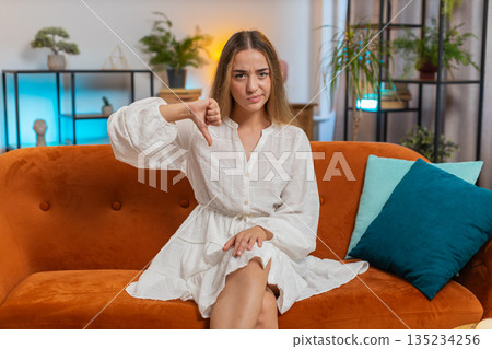 Upset young woman showing thumbs down sign gesture, expressing discontent sitting on sofa at home 135234256