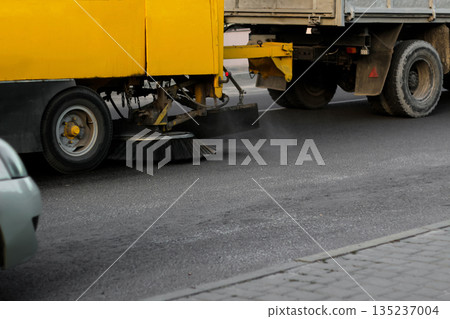 A yellow road sweeper machine is cleaning the asphalt on a city street. The circular brush is visible as it sweeps dust and debris 135237004