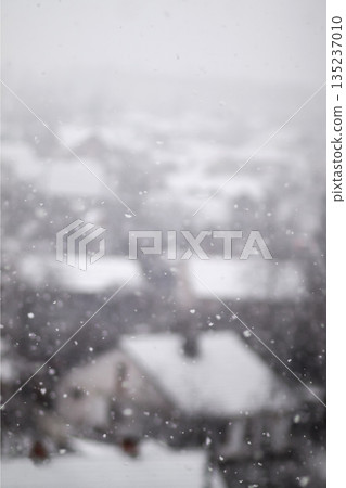 A vertical, blurry shot of snowflakes falling over a snow-covered town. The houses and roofs are out of focus in the background, creating a soft, wintry atmosphere. Vertical snowfall background A vertical, blurry shot of snowflakes falling over a snow-covered town. The houses and roofs are out of focus in the background, creating a soft, wintry atmosphere. Vertical snowfall background 135237010