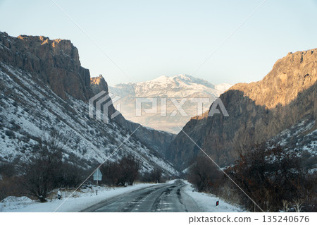Mountain road runs through a snow-covered gorge in Armenia, dramatic rocky cliffs, winter landscape, clear sky, sense of depth and journey 135240676