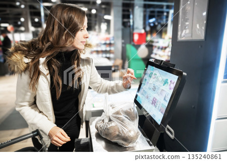 A woman stands at a self-checkout terminal in a supermarket. She is using a touchscreen to scan items, including a bag of potatoes, as customers shop around her 135240861