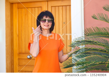 An adult woman in a short bright dress in the garden of her house near a palm tree An adult woman in a short bright dress in the garden of her house near a palm tree 135240904