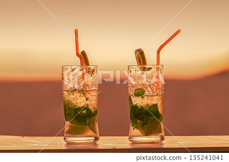 Two mojito cocktails on the background of an orange sunset. The concept of relaxing on the resort 's beach and traveling together . Honeymoon trips to warm countries. High quality photo 135241041