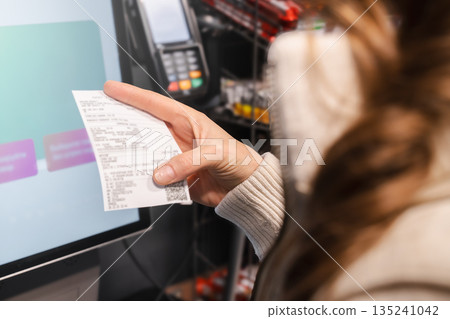 Woman examines her receipt closely at a self-service checkout while using the touchscreen display in a retail environment. Managing household expenses Woman examines her receipt closely at a self-service checkout while using the touchscreen display in a retail environment. Managing household expenses 135241042