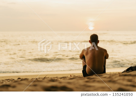 A young athletic man with a strong and powerful back sits on the sand on the beach by the sea and admires the beautiful sunset and the sun above the water. Solo travel copy space yellow background A young athletic man with a strong and powerful back sits on the sand on the beach by the sea and admires the beautiful sunset and the sun above the water. Solo travel copy space yellow background 135241050