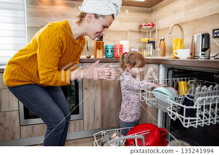 Little girl helping her mum load the dishwasher in the kitchen Little girl helping her mum load the dishwasher in the kitchen 135241994