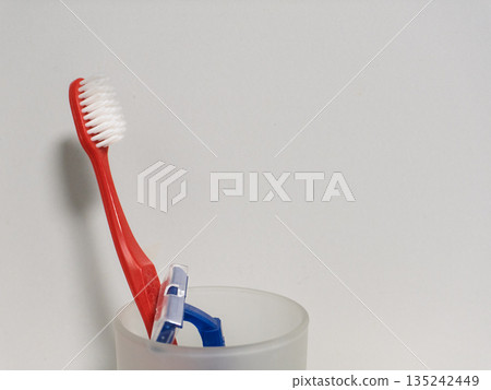Red toothbrush and blue razor in a frosted glass cup. Dental hygiene and grooming accessories in bathroom. Copy space for text 135242449