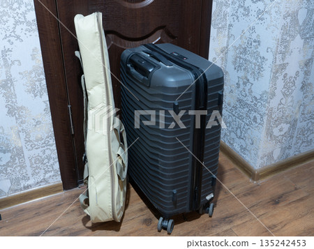 Dark grey hard-shell suitcase and beige guitar case standing by a wooden door. Luggage and musical instrument bag ready for travel or tour. Musician packing concept Dark grey hard-shell suitcase and beige guitar case standing by a wooden door. Luggage and musical instrument bag ready for travel or tour. Musician packing concept 135242453