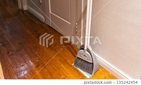 Grey broom and dustpan set standing on worn wooden floor. Domestic cleaning tools against beige wall in hallway. Housework and chores concept Grey broom and dustpan set standing on worn wooden floor. Domestic cleaning tools against beige wall in hallway. Housework and chores concept 135242454