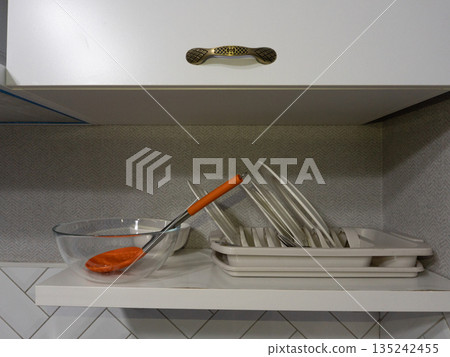 Kitchen shelf with drying rack holding white plates. Glass bowl with orange ladle under cabinet handle Kitchen shelf with drying rack holding white plates. Glass bowl with orange ladle under cabinet handle 135242455