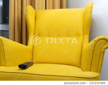 Yellow wingback armchair with a remote control on the seat. Comfortable furniture in a modern living room interior. Watching TV concept Yellow wingback armchair with a remote control on the seat. Comfortable furniture in a modern living room interior. Watching TV concept 135242460