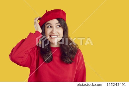 Confused smiling brunette woman in red beret and sweatshirt looking aside on yellow background. Confused smiling brunette woman in red beret and sweatshirt looking aside on yellow background. 135243491