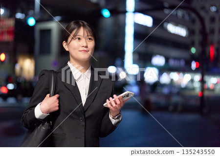 A young businesswoman standing in the city at night with a smartphone in her hand 135243550