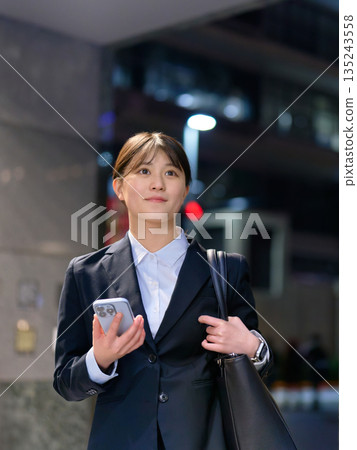 一位身穿西裝的女士下班回家，手裡拿著一支智慧型手機。 135243558