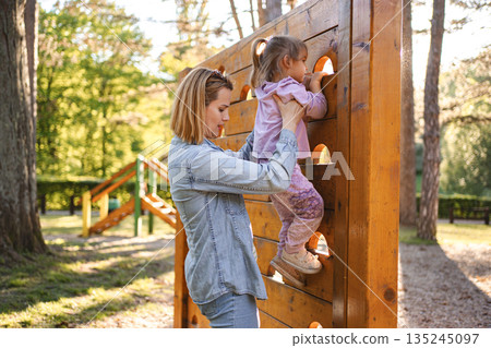 Smiling mom helps her cute daughter climb the playground Smiling mom helps her cute daughter climb the playground 135245097