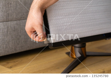 Close-up of a man's hand carefully peeling a clear protective film from the screen of a new high-performance gaming monitor during unboxing Close-up of a man's hand carefully peeling a clear protective film from the screen of a new high-performance gaming monitor during unboxing 135245780