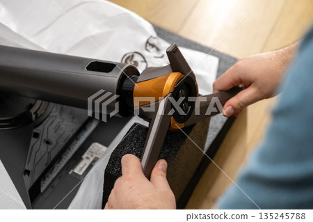 A person's hands skillfully assembling a modern monitor stand featuring sleek design, distinctive orange accents, and precise components for an optimized workspace setup 135245788