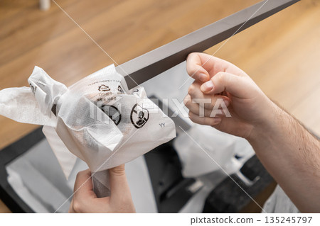 Hands carefully unboxing a new sleek silver monitor stand from protective plastic film, displaying recycling symbols and OTHER waste instructions Hands carefully unboxing a new sleek silver monitor stand from protective plastic film, displaying recycling symbols and OTHER waste instructions 135245797