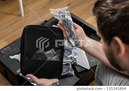 Man unboxes new electronic device accessories, holding a black zippered case and a power cable, highlighting efficient organization for technology setup Man unboxes new electronic device accessories, holding a black zippered case and a power cable, highlighting efficient organization for technology setup 135245804