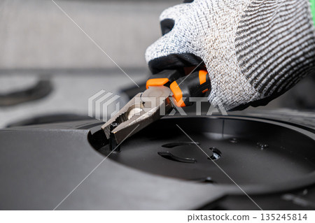 Skilled professional's gloved hand expertly uses orange-handled pliers to adjust a tiny component on a sleek black electronic device, highlighting detailed technical assembly or repair work Skilled professional's gloved hand expertly uses orange-handled pliers to adjust a tiny component on a sleek black electronic device, highlighting detailed technical assembly or repair work 135245814