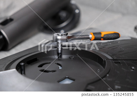 A metallic ratchet wrench with an orange handle tightening a screw on a black electronic device base, representing repair and DIY assembly 135245855