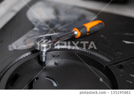 Chrome ratchet wrench with an ergonomic orange handle fastening a hex screw on a dark electronics base, showcasing precision technical repair and equipment assembly Chrome ratchet wrench with an ergonomic orange handle fastening a hex screw on a dark electronics base, showcasing precision technical repair and equipment assembly 135245861