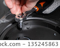 Gloved hand uses a chrome ratchet wrench to tighten a bolt on a dark mechanical device, representing precise assembly and repair work 135245863