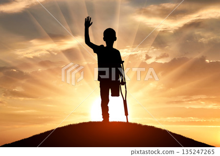 A silhouette of a young disabled boy with a crutch standing on a hill 135247265