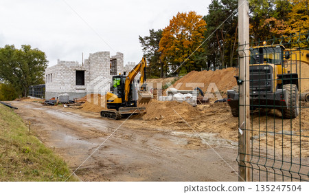 Rogachev, Belarus - October 8, 2025 - Yellow excavator against the backdrop of a brick building under construction and a pile of sand 135247504
