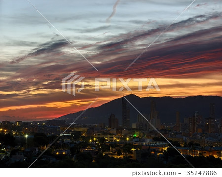 bright urban illumination contrasts with fiery sunset over jagged mountain silhouette in coastal bright urban illumination contrasts with fiery sunset over jagged mountain silhouette in coastal 135247886
