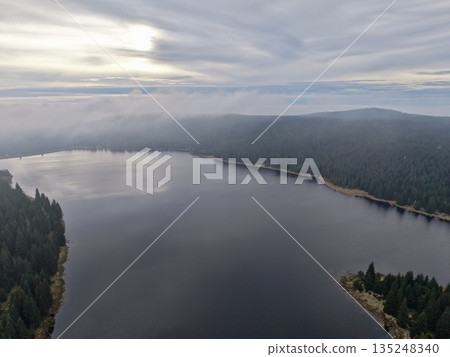 Calm Forest Lake At Sunrise With Haze, Quiet Wilderness Scene Featuring Reflections And Distant Hills 135248340