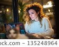 Woman works on laptop at night in cozy cafe, illuminated by warm lighting, cat nearby. 135248964