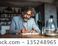 Focused mature man writing in notebook, serious expression, glasses, curly hair, grey blazer, indoor setting, thoughtful, concentrated. 135248965