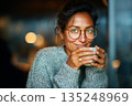 Woman enjoying warm beverage in cozy sweater sits near window in cafe, relaxing and content 135248969