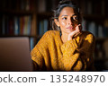 Thoughtful young woman in yellow sweater contemplates near laptop in a library setting 135248970