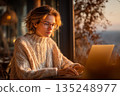 Woman working on laptop in cozy autumn cafe enjoying warm sunset light near window 135248977