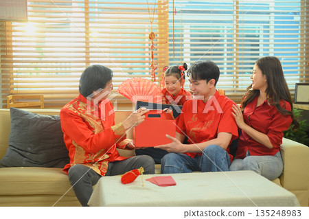 A man presents a festive box of orange to father while family gathers together during a warm Chinese New Year celebration at home. A man presents a festive box of orange to father while family gathers together during a warm Chinese New Year celebration at home. 135248983