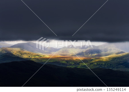 A picturesque view in Killarney National Park, Ireland. Sunlight breaks through a dark, stormy sky, illuminating the rolling green hills and valley below in a breathtaking scene. A picturesque view in Killarney National Park, Ireland. Sunlight breaks through a dark, stormy sky, illuminating the rolling green hills and valley below in a breathtaking scene. 135249114