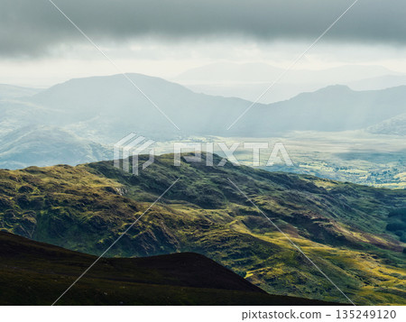 This mountainous landscape shows a valley in County Kerry, Ireland. The cloud-covered sky filters the sunlight, creating rays that shine down on the green land. Mountains rise in the background. 135249120