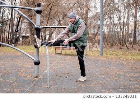 A man in a tracksuit and hat stretches on an outdoor sports ground. Exercising in cold weather. 135249331