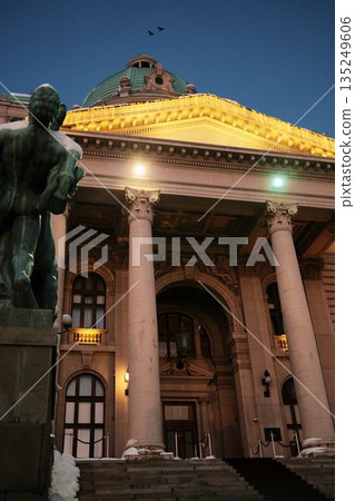 Monument stands before an illuminated historic building during winter evening. The scene represents cultural heritage, urban identity, and seasonal city atmosphere 135249606