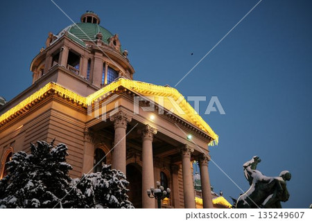 Monument stands before an illuminated historic building during winter evening. The scene represents cultural heritage, urban identity, and seasonal city atmosphere Monument stands before an illuminated historic building during winter evening. The scene represents cultural heritage, urban identity, and seasonal city atmosphere 135249607
