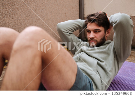 A man exercises on a mat doing abdominal crunches. The scene shows a focused wellness routine at home 135249668
