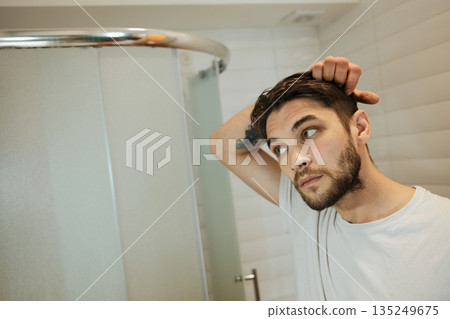 A man examines his hair in front of a bathroom mirror. The scene highlights personal care and a simple daily ritual A man examines his hair in front of a bathroom mirror. The scene highlights personal care and a simple daily ritual 135249675