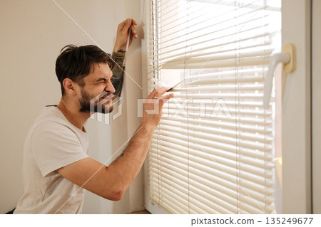 A man peeks through window blinds with a playful squint. The moment reflects curiosity and a light everyday routine 135249677