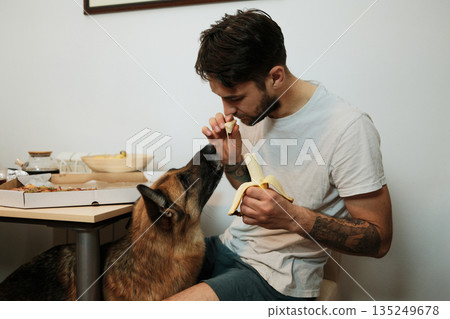A man eats a banana at the table while offering a small piece to a dog. The moment shows trust, playfulness, and everyday living 135249678