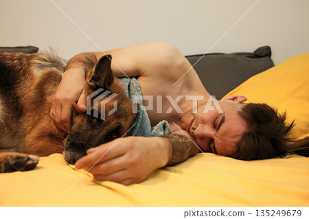 A man sleeps on a bed hugging a dog under a blanket. The moment captures tenderness and quiet emotional warmth A man sleeps on a bed hugging a dog under a blanket. The moment captures tenderness and quiet emotional warmth 135249679