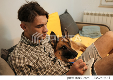 A man writes in a notebook while sitting on a couch with a dog resting close. The scene reflects calm routine and quiet connection at home A man writes in a notebook while sitting on a couch with a dog resting close. The scene reflects calm routine and quiet connection at home 135249681