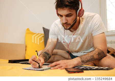 A man leans over a notebook and writes while wearing headphones, with a roll-up keyboard spread out on the bed. The scene shows focused music planning and the concept of creative work 135249684